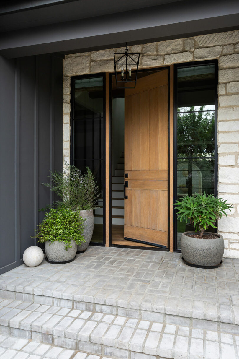 Wooden front door slightly ajar, flanked by plants in pots on a brick porch. Steps lead up to the entrance with a lantern overhead.