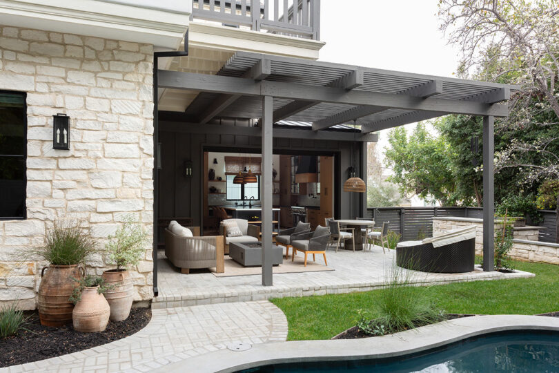 Patio al aire libre con una pérgola de madera oscura, asientos beige, una mesa de comedor, plantas en macetas y una piscina en primer plano. El patio se abre en una casa con grandes ventanas y muros de piedra.