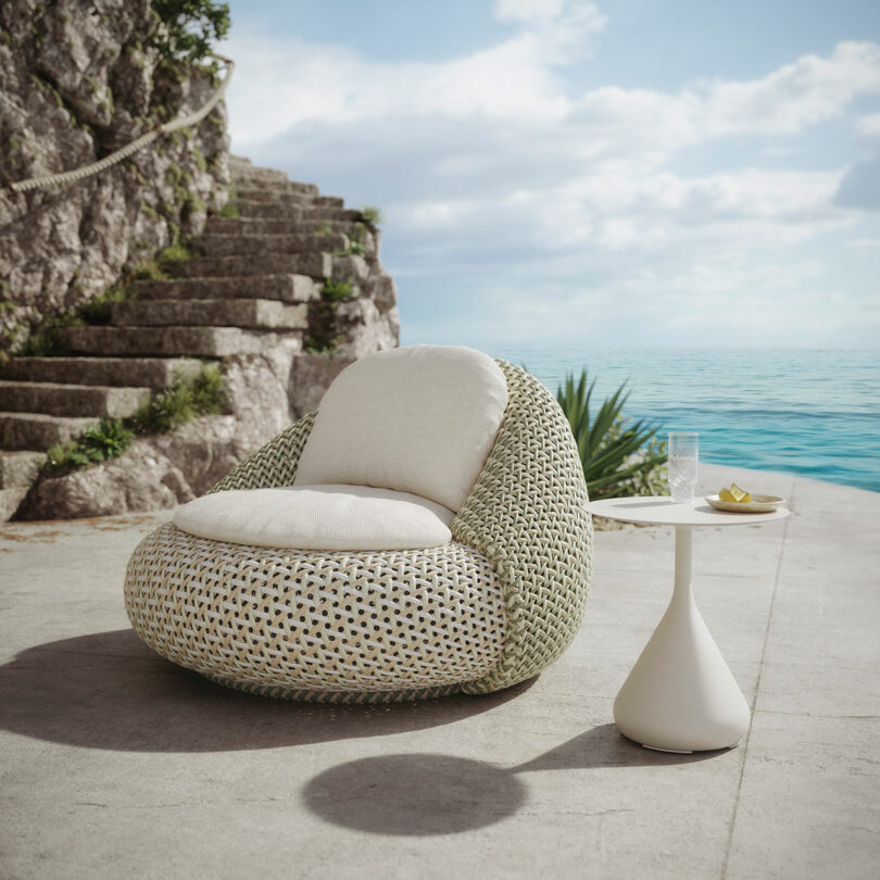 An exquisite outdoor scene featuring a DEDON wicker armchair and a small white table on a stone patio overlooking the ocean, with charming stone steps and lush greenery enhancing the backdrop.