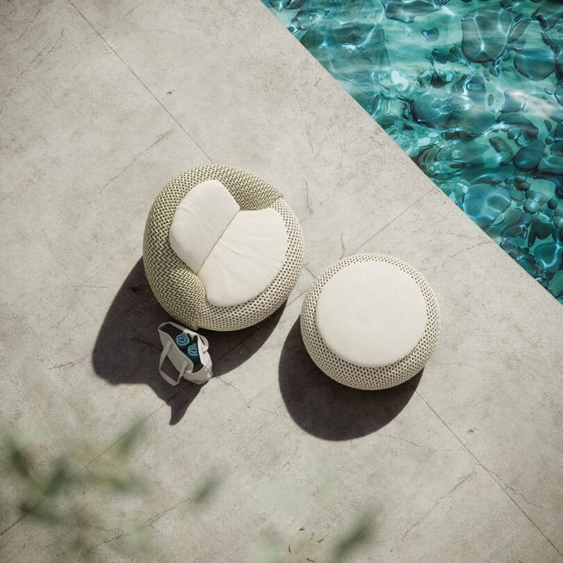 Two round beige DEDON chairs sit serenely on a concrete poolside, accompanied by a stylish tote bag. The crystal-clear blue water of the pool is just partially visible, offering an inviting glimpse into relaxation.