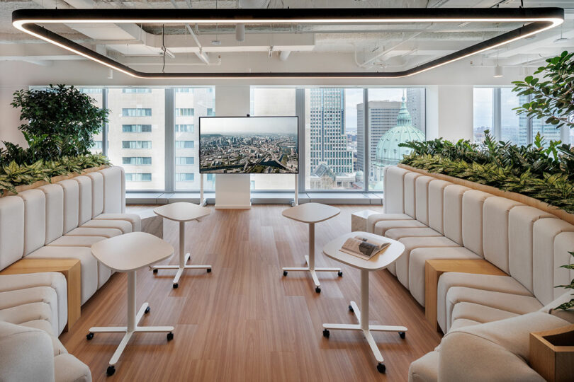 Modern conference room with curved seating, small mobile tables, and a large screen displaying a city view. Surrounded by green plants, the room has large windows showing urban buildings outside.