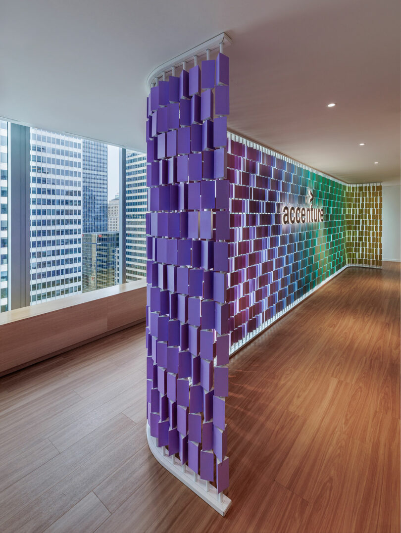 Office lobby with a colorful pixelated wall display and wooden floor. Large windows reveal a cityscape outside.