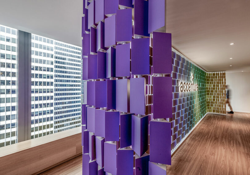 Office interior with a wall of purple rectangular panels, large windows showing skyscrapers, wooden floors, and a blurred figure walking near the "accenture" logo on the wall.