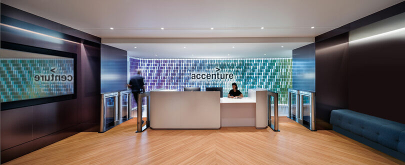 A modern office reception area with a central desk, a person sitting behind it, and a large "accenture" sign on a tiled wall.