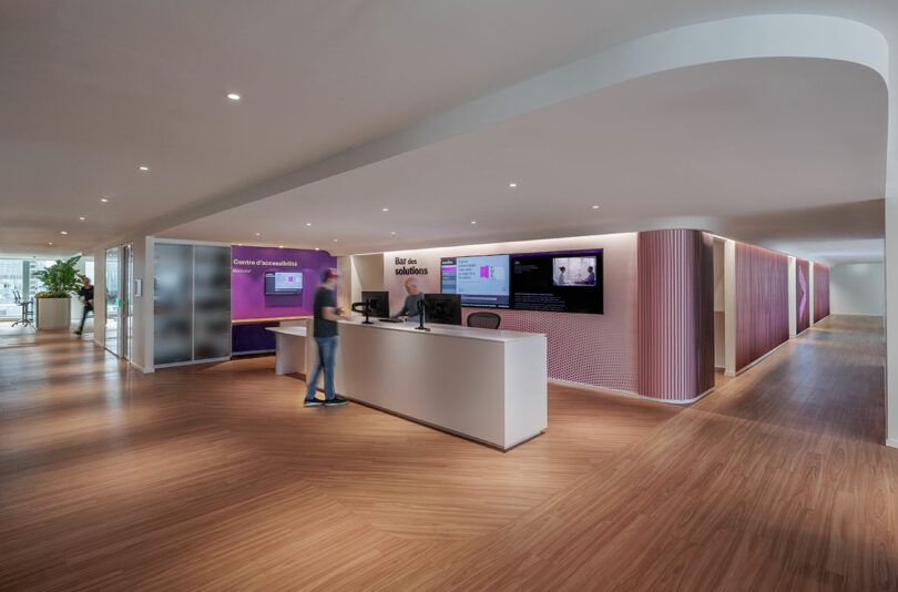 Modern office reception area with a curved white desk, two monitors, and wooden flooring. A person stands at the desk in an open, spacious environment with purple and white accents.