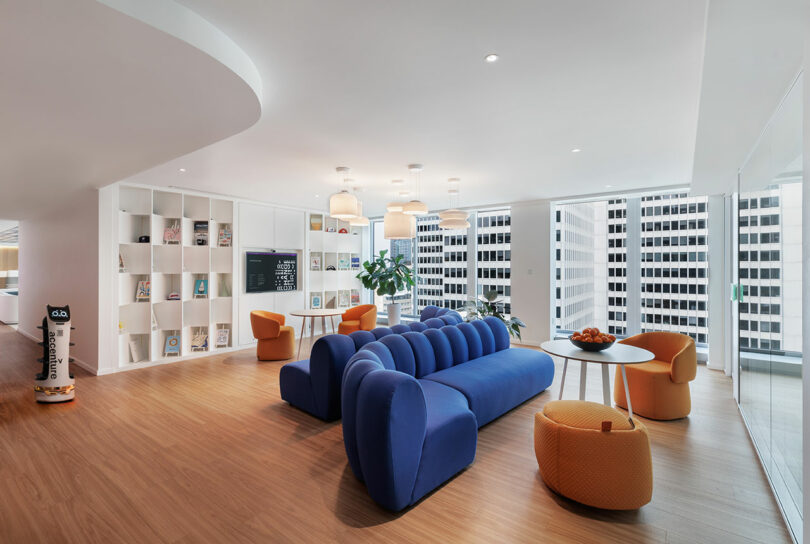 Modern office lounge with a blue sofa, orange chairs, small tables, and a bookshelf. Large windows offer a city view, and the space features wooden floors and ceiling lights.