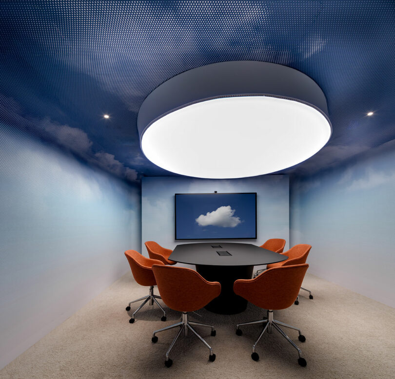 A modern meeting room features a large white circular light above a black table, surrounded by orange chairs. The walls have a blue sky pattern, and a screen displays a cloud image.