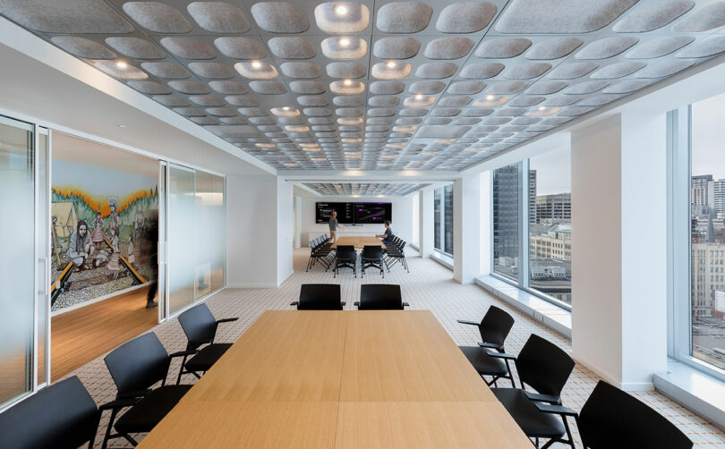 Modern conference room with long wooden table, black chairs, and large windows showing city view. Ceiling features grid pattern lighting. Mural on wall in adjacent room.