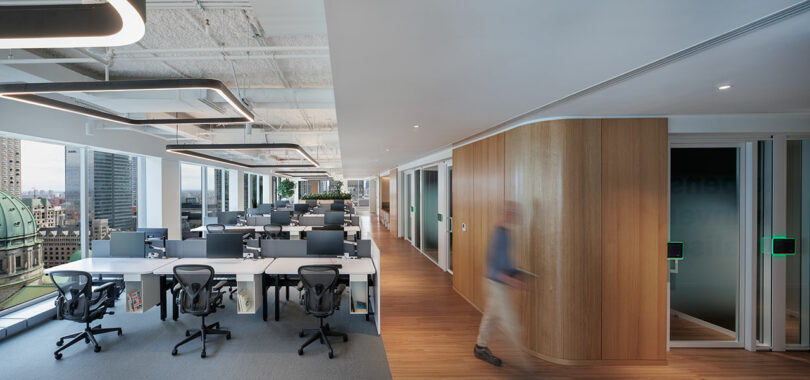 Modern office space with rows of desks and chairs, large windows, and wood-paneled walls. A person walks by on hardwood flooring, with city buildings visible outside.