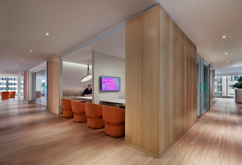 Modern office interior with wood paneling, light wood flooring, and orange chairs. A man is seated at a meeting booth with a wall-mounted screen displaying a purple interface.