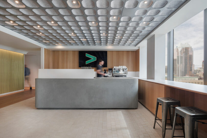 Modern office lobby with a concrete reception desk, a blurred figure behind it, wooden accents, a logo on the wall, tall windows showing cityscape, and two black stools in the foreground.