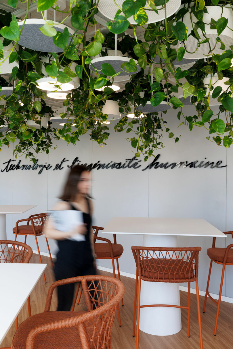 A woman walks past high tables and chairs in a modern office space with hanging plants and a wall sign reading "technologie et humanité.