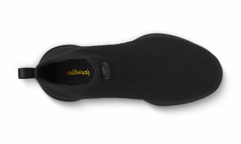 Top view of a black slip-on shoe with a ribbed fabric texture and a loop at the back. The word "allbirds" is visible inside.