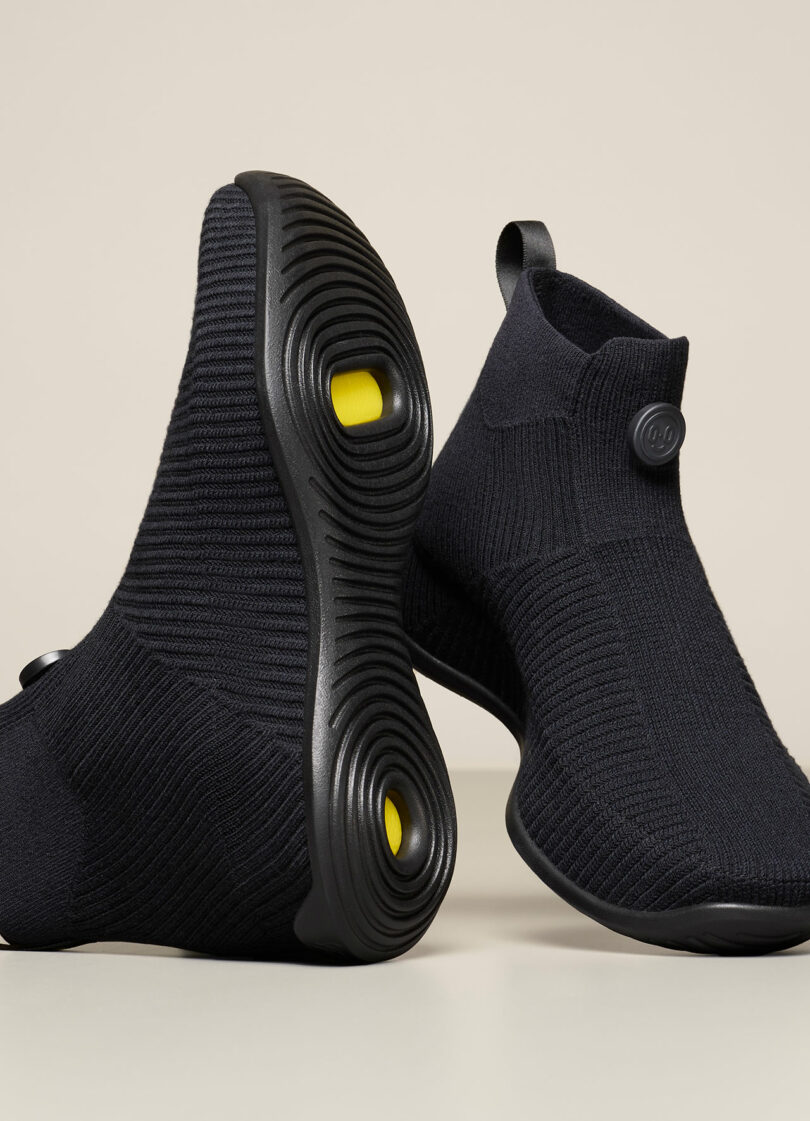 Black knitted sneakers with ribbed texture, featuring yellow accents on the sole and a button detail on the upper part.