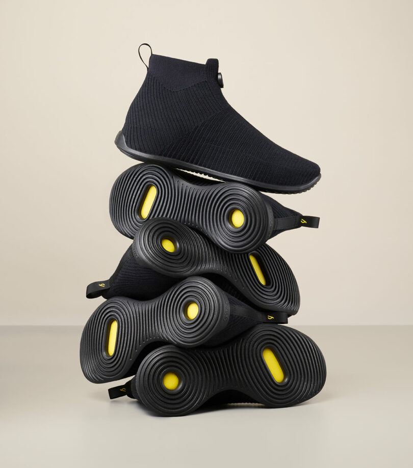 Stack of five black ankle-high shoes with circular yellow accents on the soles, placed against a neutral background.