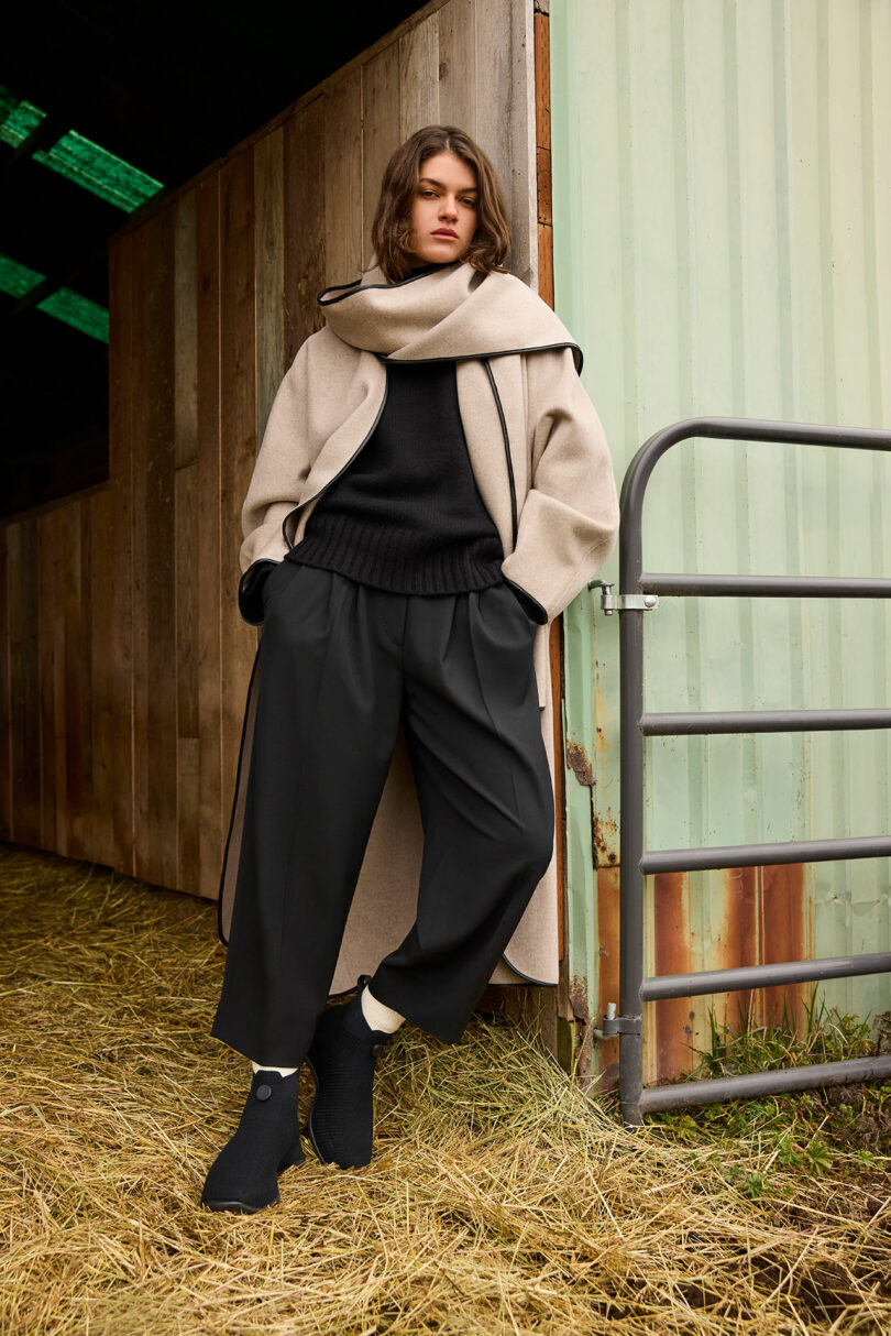 Person wearing a long beige coat and black attire stands in a barn doorway on hay-covered ground.