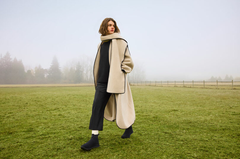 Person in a long beige coat and black boots stands on a grassy field with a foggy background.