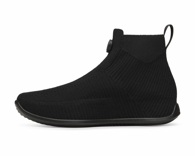 A black, ankle-high, sock-like shoe with a round toe and ribbed texture, featuring a pull tab at the heel.