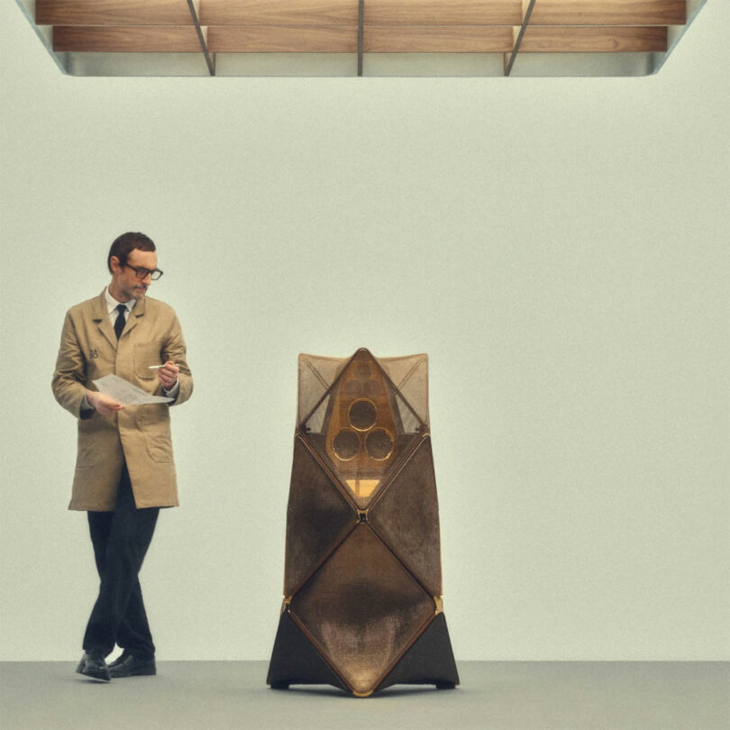 A man in a beige coat examines a Bang & Olufsen device with speaker-like elements in a minimalist room.