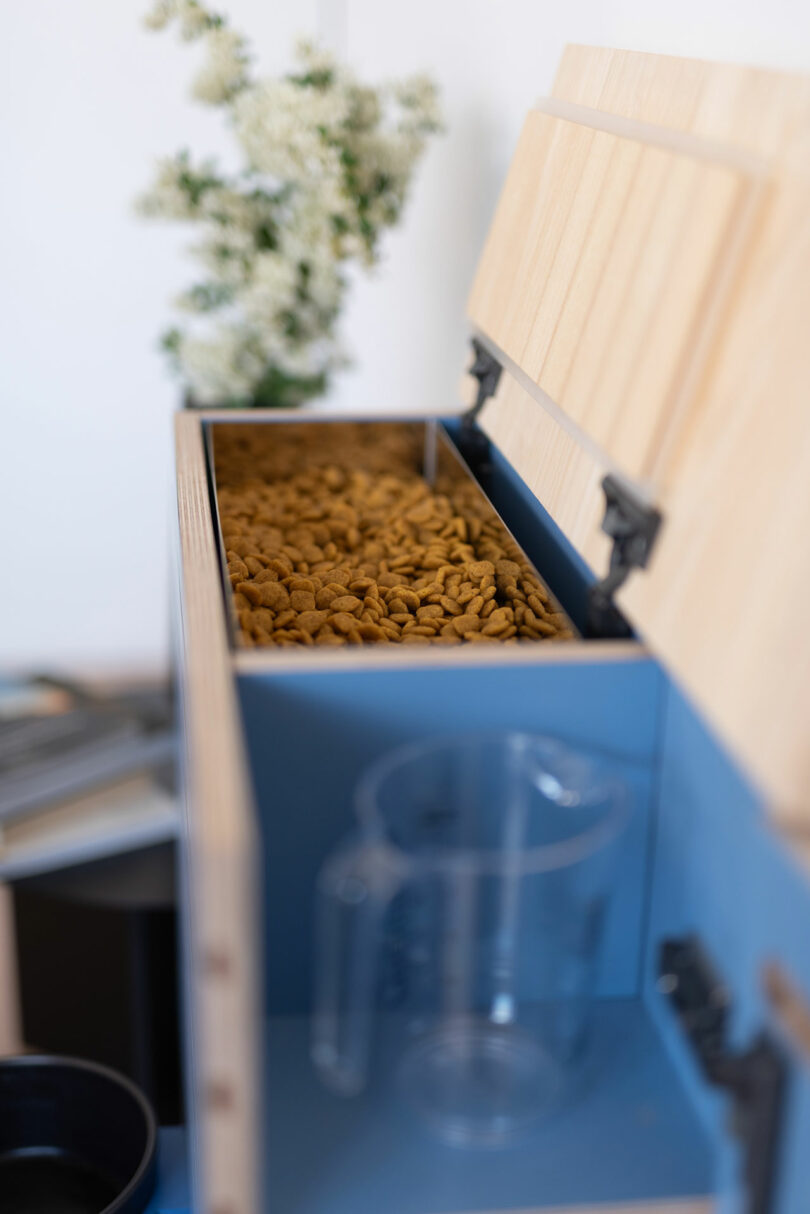 An open wooden storage box filled with dry pet food, showcasing its fresh, biting edge. A clear measuring container stands ready in front of the box, while white flowers are blurred softly in the background.