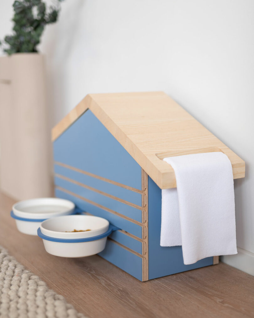 A biting edge design, this blue and wooden pet feeding station is shaped like a house with two ceramic bowls and a cloth draped over the roof.