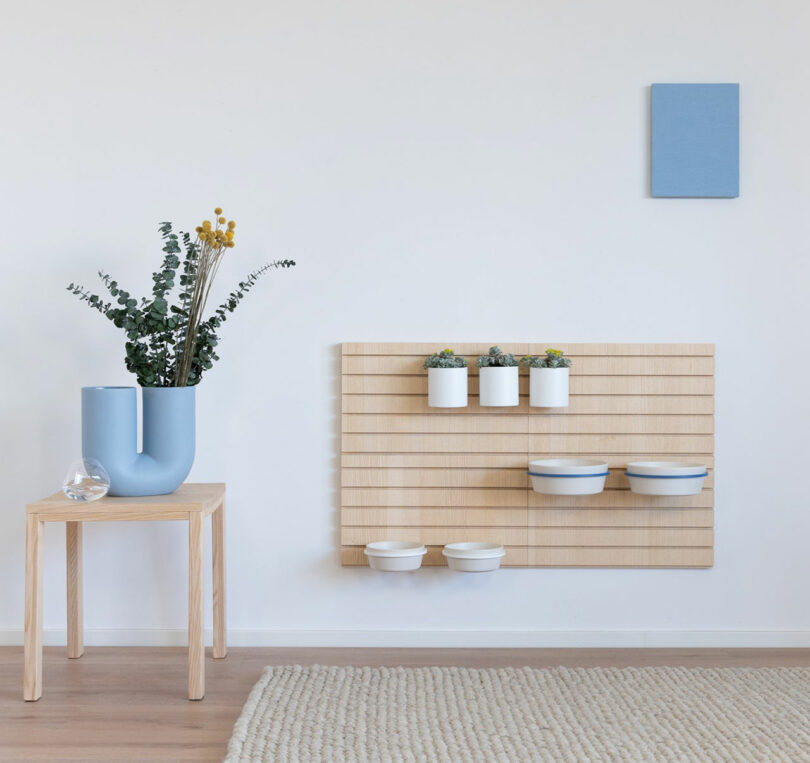 A minimalist room featuring a wooden stool with a blue vase and greenery exudes a biting edge. A wall-mounted wooden organizer with white pots and a bold blue square canvas complement the composition, while a beige rug softens the space.