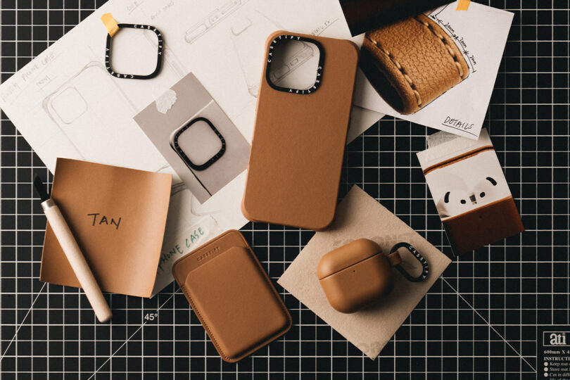 Brown leather phone case, AirPods case, and watch cover on a black grid mat with sketches and swatches.