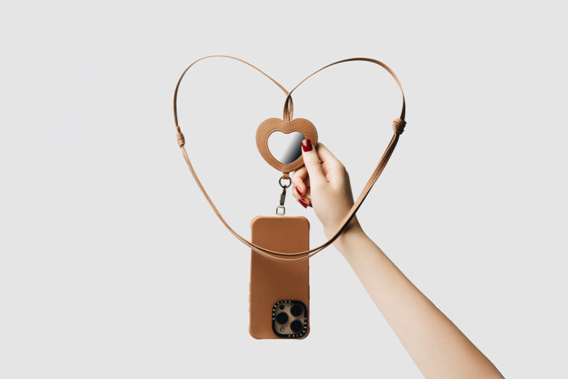 A hand holding a phone case with a strap shaped into a heart. The case is tan and the strap includes a heart-shaped loop. The phone camera lenses are visible.