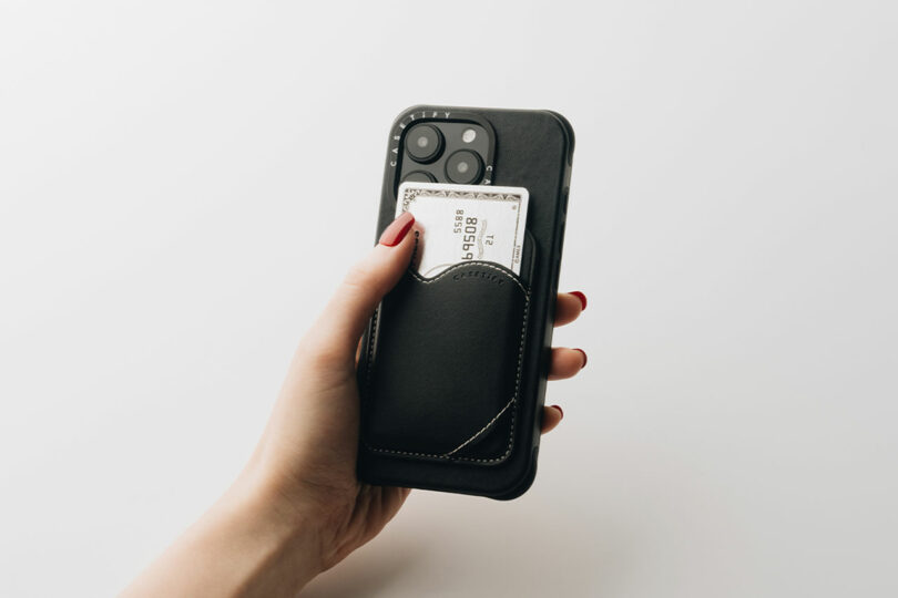 A hand grips a smartphone adorned with a sleek CASETiFY black leather card holder on the back, showcasing a glimpse of a silver card tucked inside.