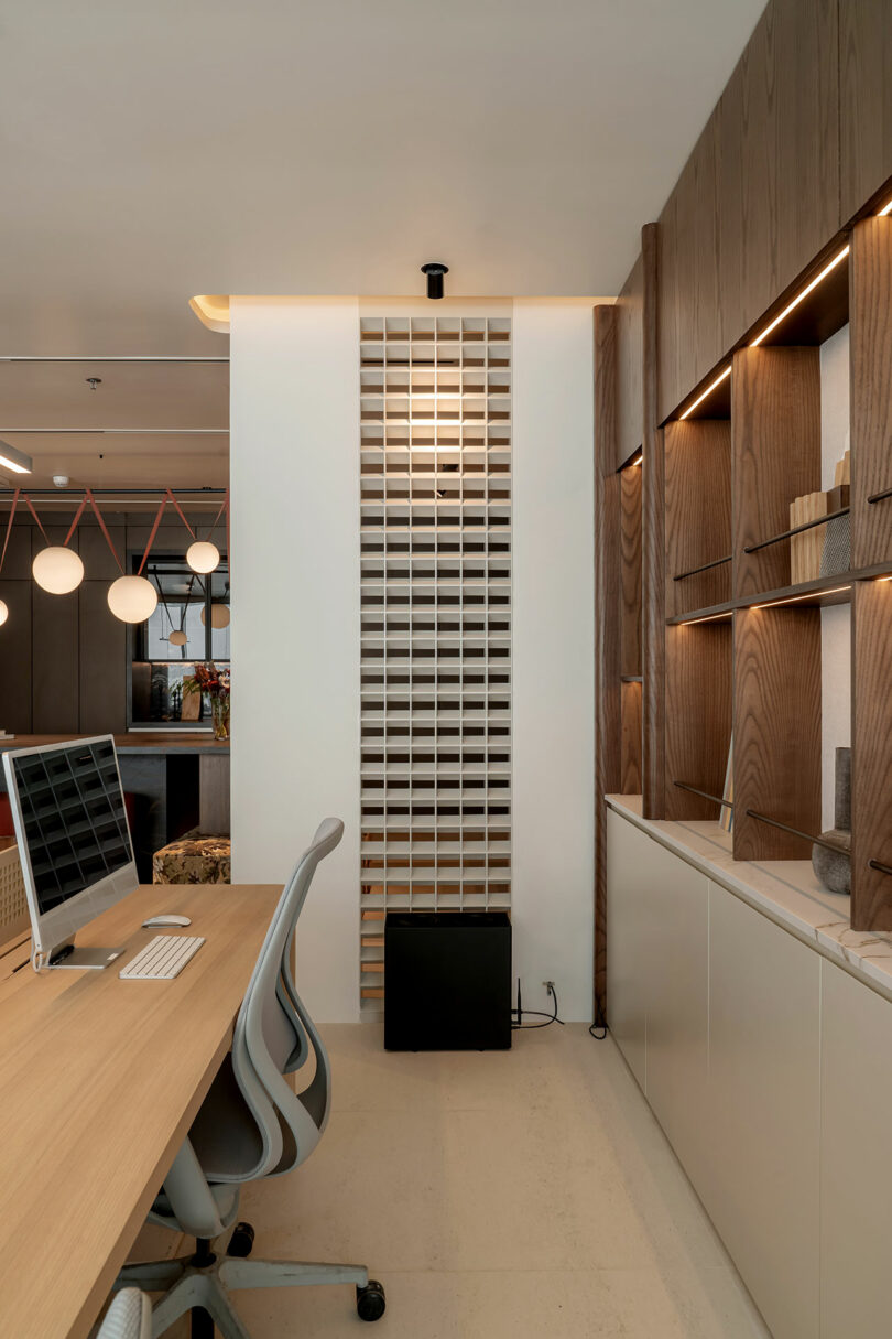 Modern office space with wooden desks, ergonomic chairs, a partition wall, and built-in wooden shelves. A computer monitor is placed on one of the desks.