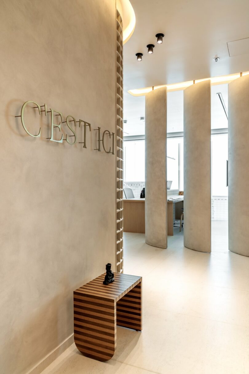 Modern interior with a beige color scheme. "C'est Ici" is displayed on a wall. A wooden bench with a small sculpture is in the foreground, and curved partition walls lead to an office space.