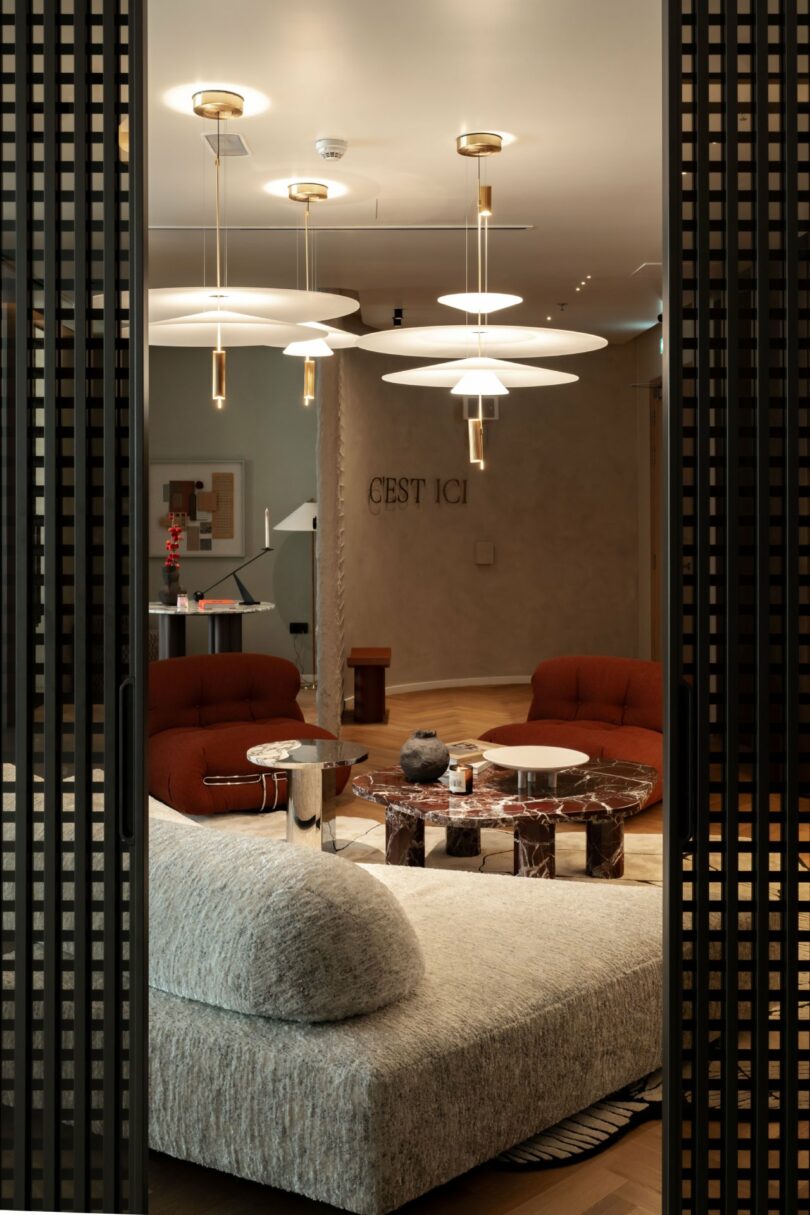 Modern lounge area with unique light fixtures, red chairs, a gray sofa, and a marble table. "Cest Ici" is written on the wall. Contemporary decor and muted color palette.