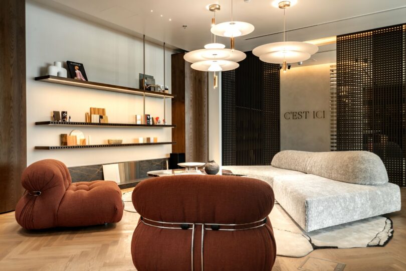 Modern living room with a gray sofa, brown chairs, two shelves holding books and decor, layered round coffee tables, pendant lights, and "C'EST ICI" on the wall.