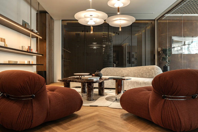 Modern living room with two brown armchairs, a textured sofa, and round tables. Pendant lights hang overhead, and a perforated wall divider is in the background. Neutral color palette.