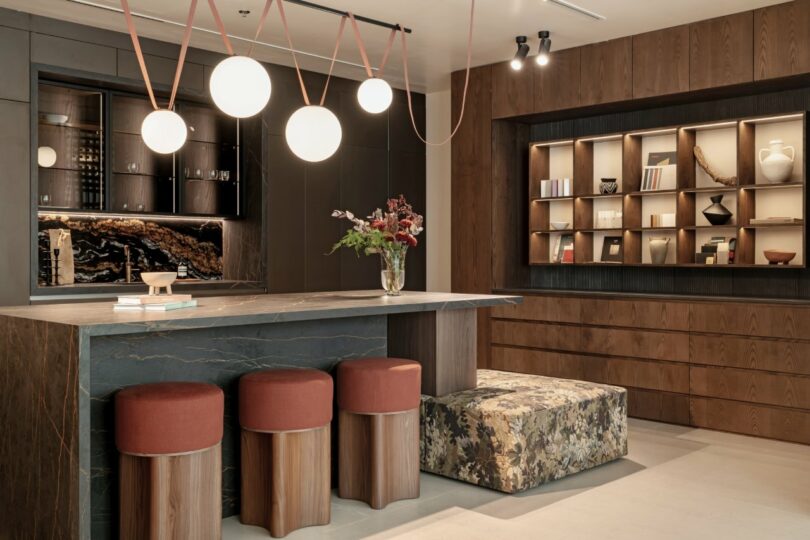 Modern kitchen with a stone island, red stools, and pendant lights. Shelving displays decor items on a wood-paneled wall, and a floral cushioned bench is nearby.