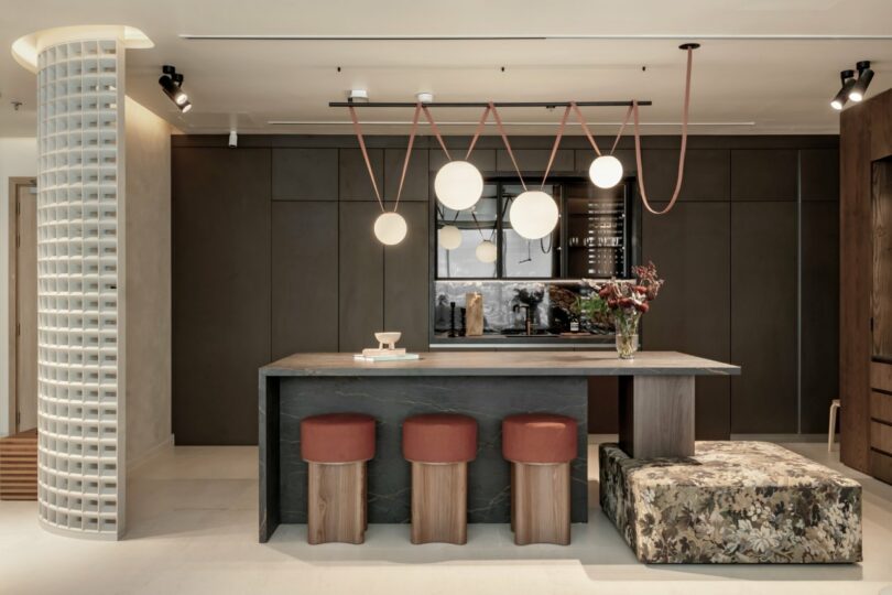 Modern kitchen with a central island, three red stools, unique pendant lighting, and a floral cushioned bench. Dark cabinetry and a circular column enhance the contemporary design.