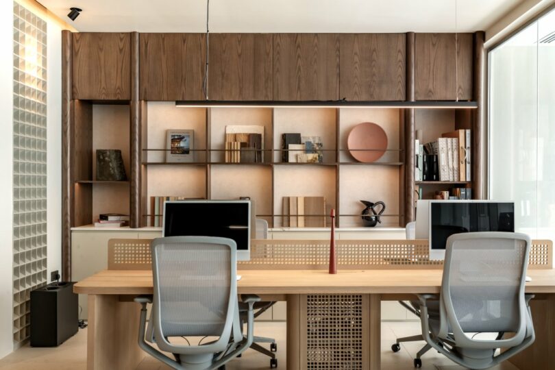 Modern office space with two desks and chairs, computers, wood shelves with books and decor, and large windows for natural light.