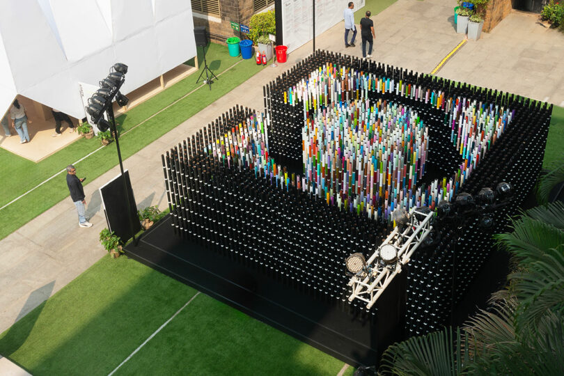 Aerial view of a vibrant Chromacosm art installation by Suchi Reddy on a black platform, surrounded by lush green spaces and walkways. Stage lights accentuate the structure's vivid colors as people gather nearby to admire its beauty.