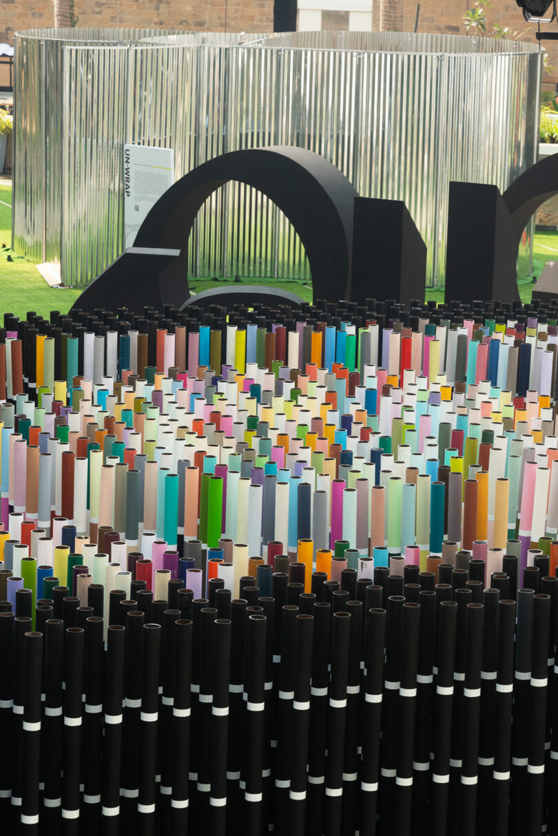 Aerial view of a vibrant Chromacosm art installation by Suchi Reddy on a black platform, surrounded by lush green spaces and walkways. Stage lights accentuate the structure's vivid colors as people gather nearby to admire its beauty.