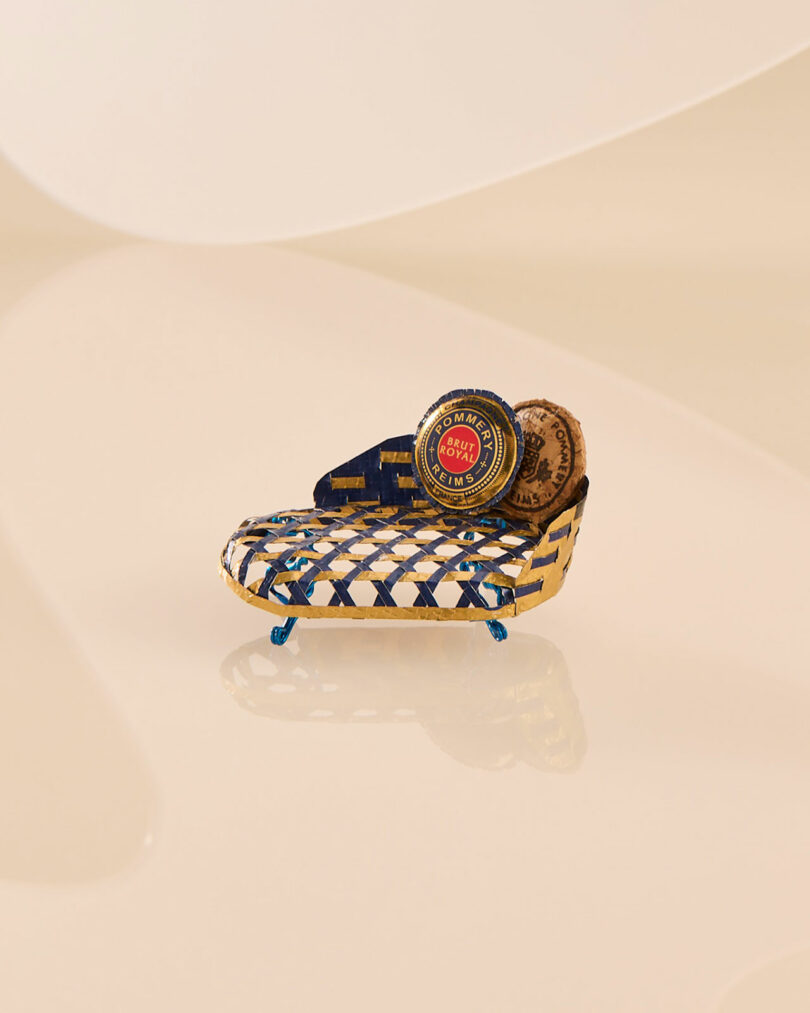 A small, intricately designed chaise longue with a patterned seat and circular pillow. The design features a blue and gold color scheme, similar to a Champagne Chair's elegance, highlighted by a decorative circular emblem on the pillow.