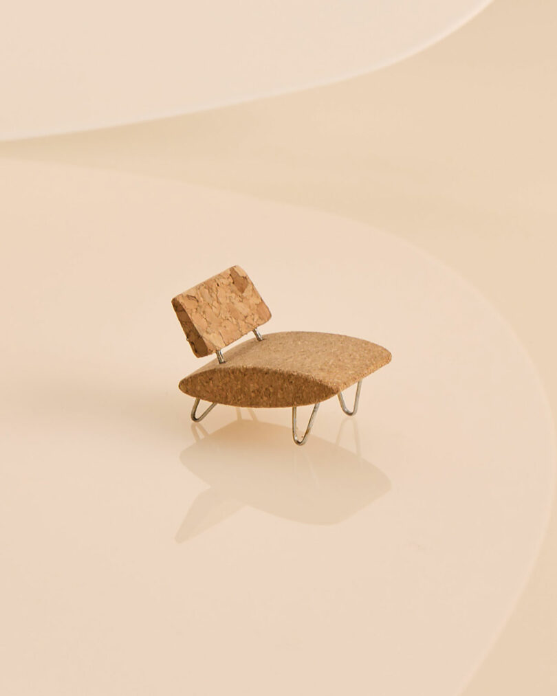 A miniature champagne chair made of cork and metal sits on a smooth, light beige surface, featuring a simple design with a curved seat and backrest.