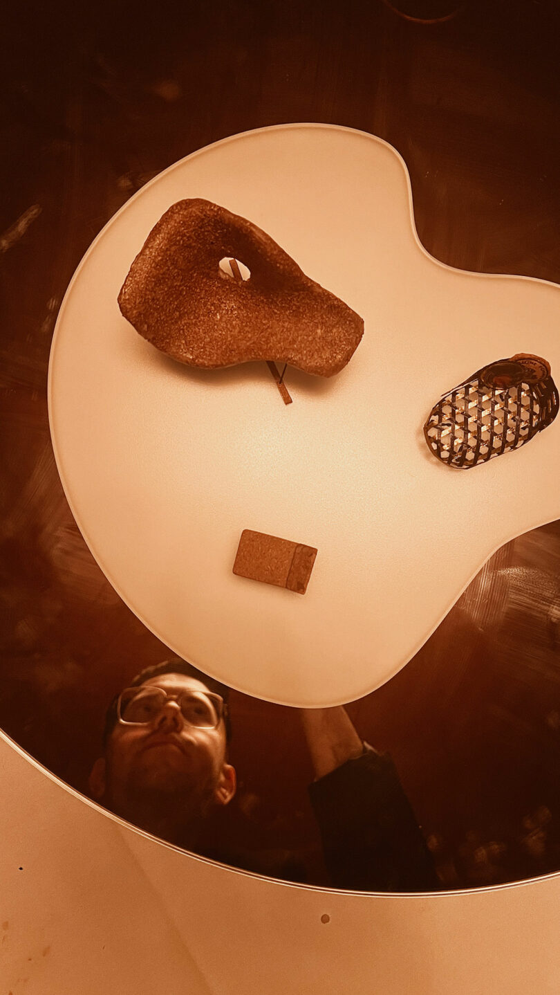 A pear, a woven object, and a small rectangular block are artfully arranged on a white surface resembling a guitar pick, akin to how elements might be balanced on a Champagne Chair. A person's reflection shimmers below.