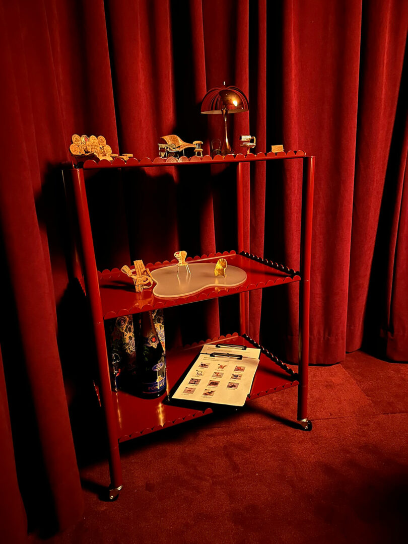 A red metal cart with three shelves hosts small decorative items, a cylindrical container, and a clipboard with papers, all watched over by plush red curtains. Nestled at the side is a stylish champagne chair, adding an elegant touch to the overall setting.