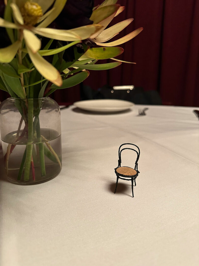 A small Champagne Chair decoratively sits on a white tablecloth beside a vase with flowers, adding a touch of elegance. A dinner plate is visible in the background.