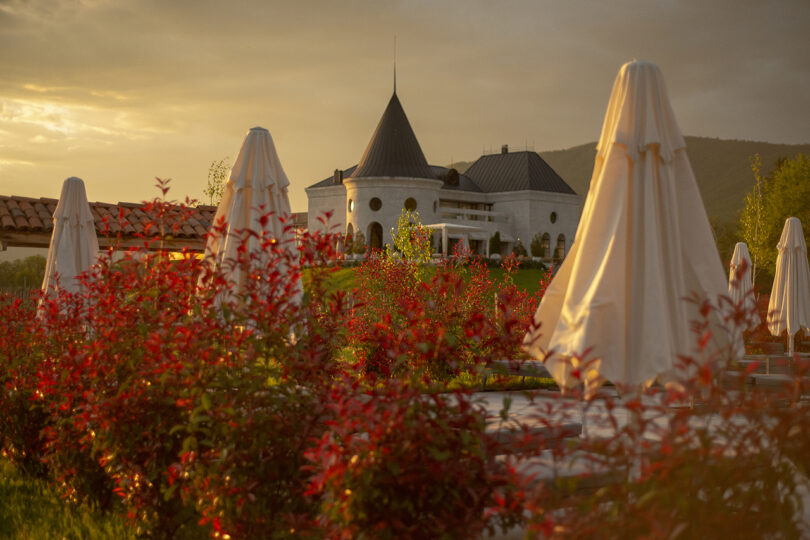 At sunset, the stone building with conical roofs at Lopota Lake Resort is enveloped by red-leaved plants and closed umbrellas.