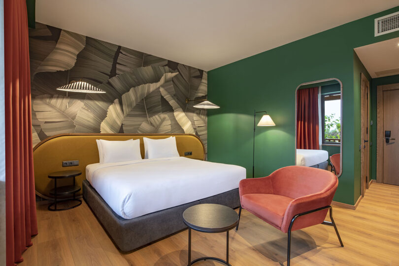 The modern hotel room at Lopota Lake Resort features green walls, leaf-patterned wallpaper, a double bed, and contemporary furnishings including a round table, red armchair, and a large mirror.