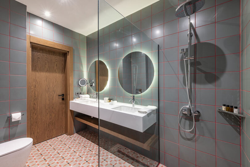 Modern bathroom at Lopota Lake Resort features gray tiled walls, a double sink with circular mirrors, and a walk-in shower. A wooden door complements the geometric-patterned floor, while amenities are neatly arranged on the counter for your convenience.