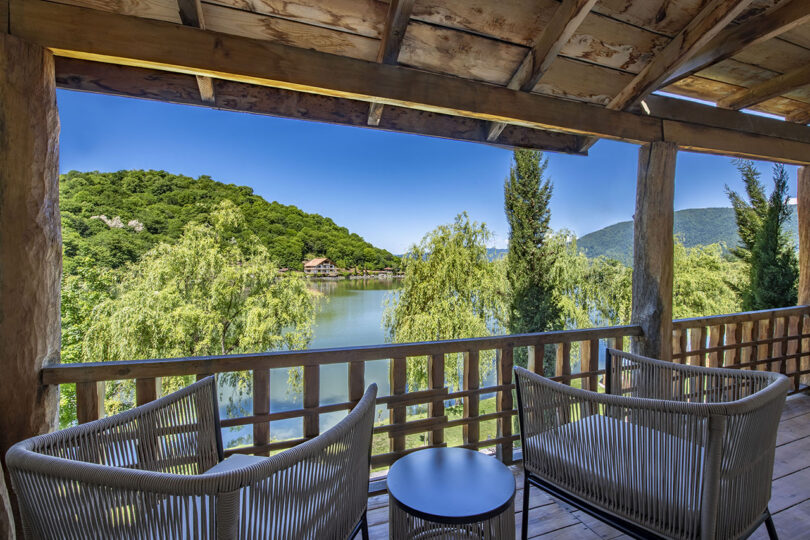 Two wicker chairs and a small table on a wooden balcony offer serene views at Lopota Lake Resort, overlooking a lake surrounded by lush green hills under a clear blue sky.