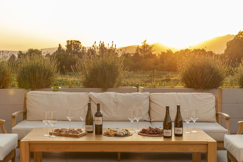 At Lopota Lake Resort, the outdoor seating area features a wooden table adorned with wine bottles, glasses, and appetizers. As the sun sets over distant hills, its warm light bathes the lavender plants in a golden hue.