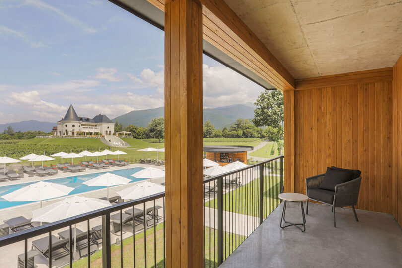 View from a wooden balcony at Lopota Lake Resort, overlooking a pool area with umbrellas and a distant castle-like building against a backdrop of mountains and blue sky.